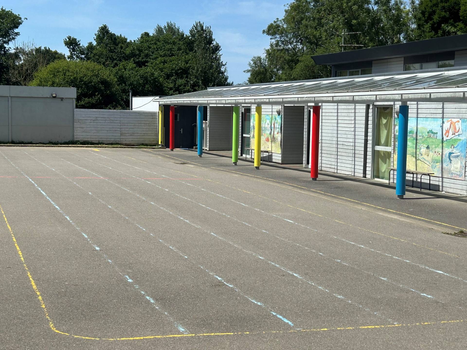 Empty school courtyard with a row of multicolored pillars beside a mural-covered building under a blue sky